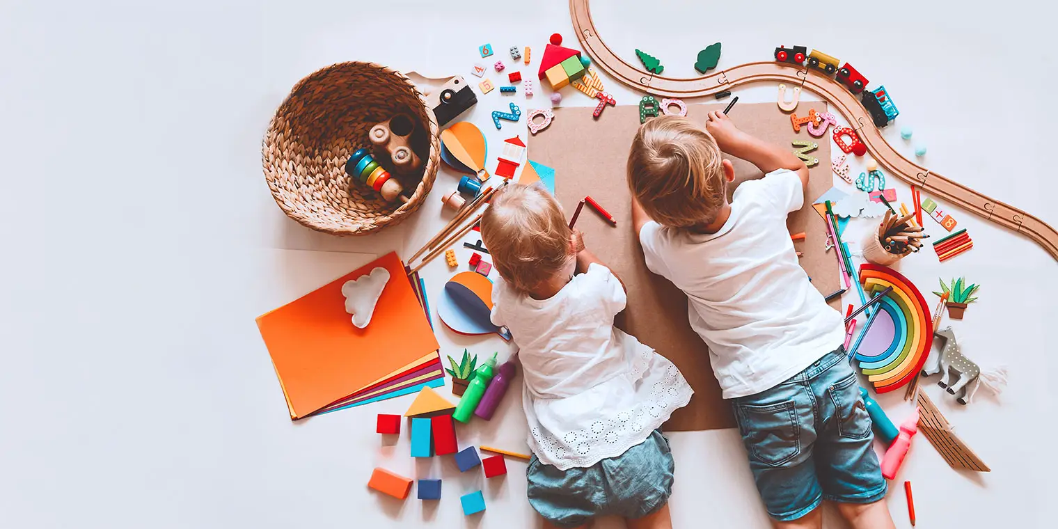 Zwei Kinder liegen auf dem Boden und zeichnen auf einem großen Blatt, umgeben von bunten Spielzeugen und Bastelmaterialien.
