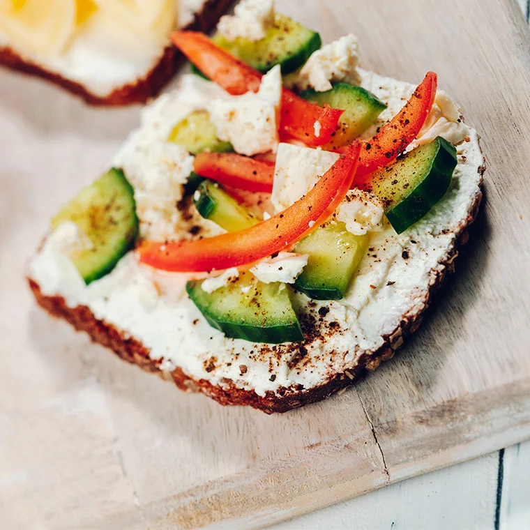 Ein belegtes Vollkornbrot mit Frischkäse, Gurke, Paprika und Gewürzen auf einem Holzbrett.