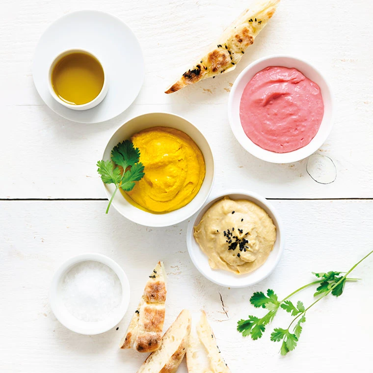 Drei verschiedene Dips in Schalen mit Fladenbrotstücken und Kräutern auf einem weißen Tisch.