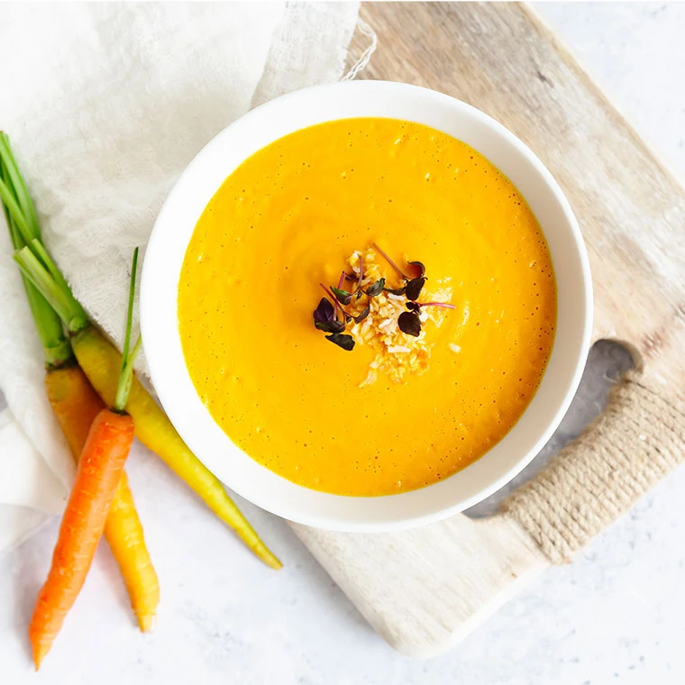 Eine Schale cremige Karottensuppe auf einem Holzbrett mit frischen Karotten daneben.