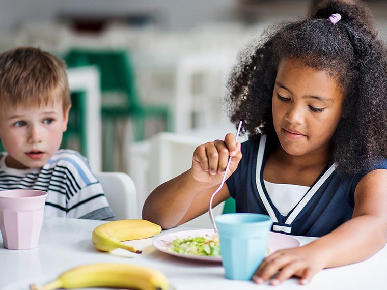 Ein Mädchen mit lockigem Haar isst konzentriert ihr Mittagessen in der Schulkantine, während ein Junge neben ihr sitzt und sich umsieht.