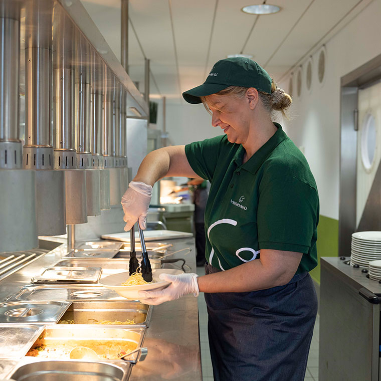 Eine Servicekraft von VielfaltMenü serviert Nudeln an einer Essensausgabe.