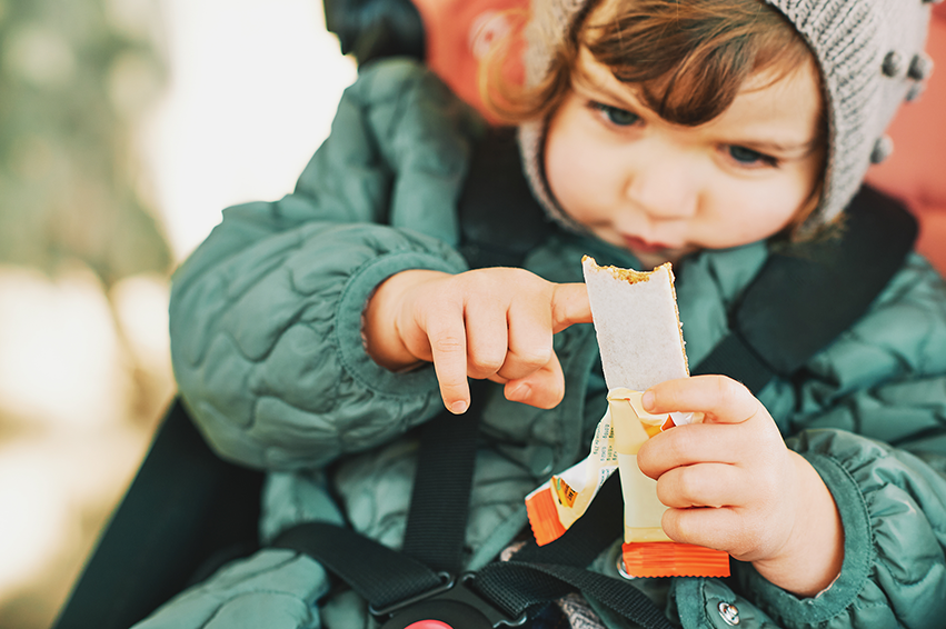 Kleinkind im Kindersitz hält einen Früchteriegel und zeigt auf die Verpackung