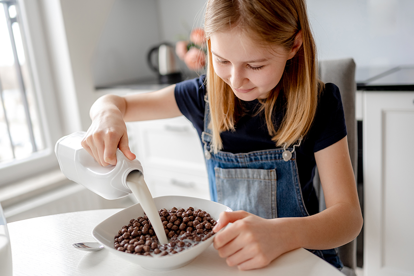 Schulkind gießt Milch in eine Schale mit Frühstückscerealien am Küchentisch.